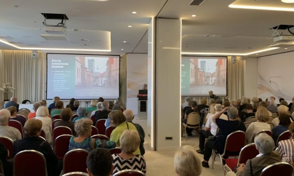 Na fotografiji se vidi dvorana ispunjena ljudima koji sjede i prate predavanje ili prezentaciju. Publika je okrenuta prema pozornici, gdje se na dva velika platna projicira isti sadržaj – prikaz ulice s tekstom na hrvatskom jeziku. U prostoriji je postavljeno više redova crvenih stolica, a atmosfera djeluje ozbiljno i usmjereno na edukativni ili informativni sadržaj. Rasvjeta je moderna i svijetla, a prostor izgleda uredno i suvremeno, s bijelim zidovima i zavjesama.