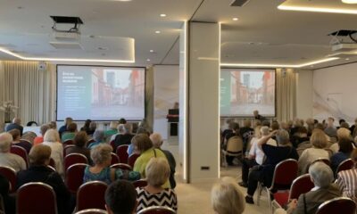 Na fotografiji se vidi dvorana ispunjena ljudima koji sjede i prate predavanje ili prezentaciju. Publika je okrenuta prema pozornici, gdje se na dva velika platna projicira isti sadržaj – prikaz ulice s tekstom na hrvatskom jeziku. U prostoriji je postavljeno više redova crvenih stolica, a atmosfera djeluje ozbiljno i usmjereno na edukativni ili informativni sadržaj. Rasvjeta je moderna i svijetla, a prostor izgleda uredno i suvremeno, s bijelim zidovima i zavjesama.