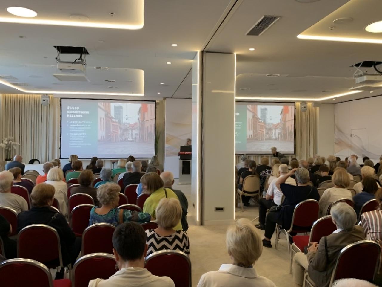 Na fotografiji se vidi dvorana ispunjena ljudima koji sjede i prate predavanje ili prezentaciju. Publika je okrenuta prema pozornici, gdje se na dva velika platna projicira isti sadržaj – prikaz ulice s tekstom na hrvatskom jeziku. U prostoriji je postavljeno više redova crvenih stolica, a atmosfera djeluje ozbiljno i usmjereno na edukativni ili informativni sadržaj. Rasvjeta je moderna i svijetla, a prostor izgleda uredno i suvremeno, s bijelim zidovima i zavjesama.