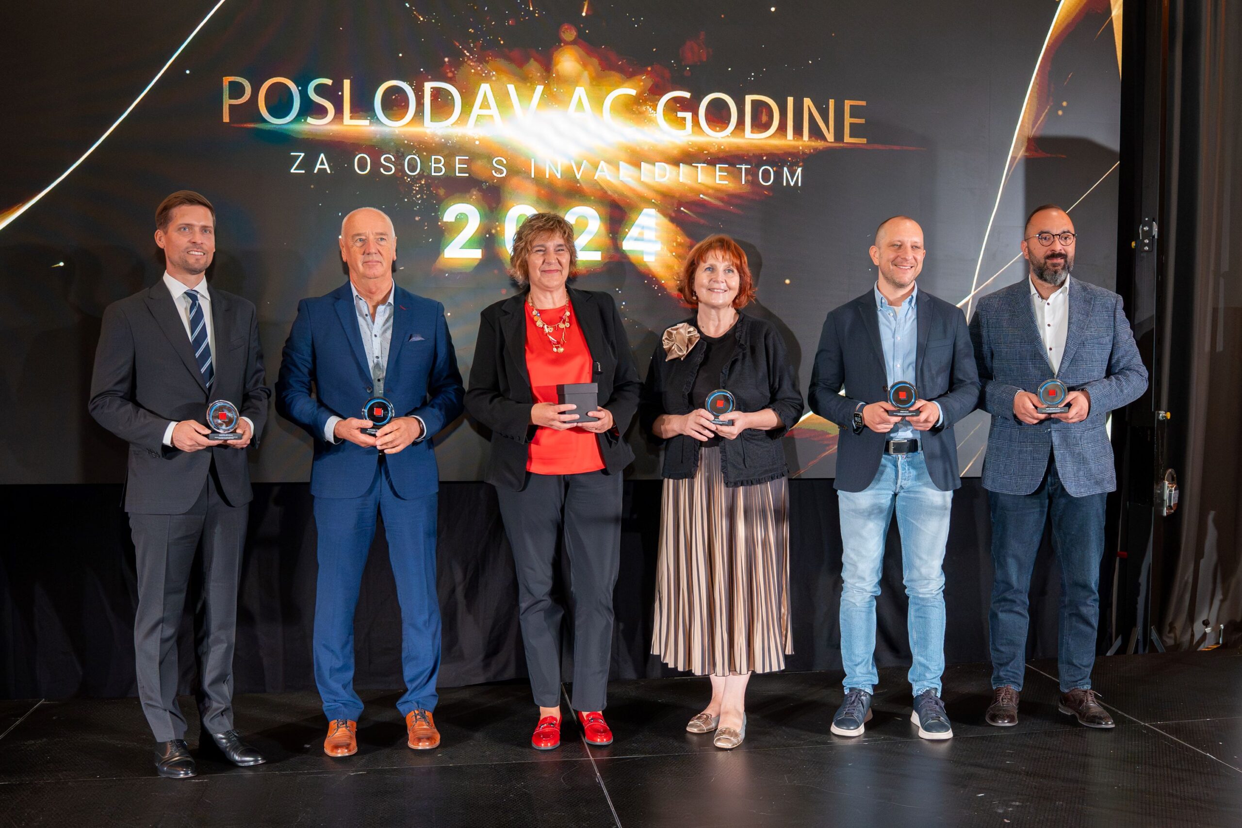 Na fotografiji se nalazi šest osoba koje stoje u nizu na pozornici, ispred velikog ekrana na kojem piše "POSLODAVAC GODINE ZA OSOBE S INVALIDITETOM 2024". Svi drže nagrade u rukama i nasmijani su. Riječ je o svečanoj dodjeli priznanja, a pozadina je tamna s efektnim svjetlosnim detaljima u zlatnoj i narančastoj boji. Osobe su svečano odjevene – muškarci u odijelima ili sakou s košuljom, a žene u elegantnoj odjeći. Atmosfera odiše svečanošću i priznanjem za postignuća.