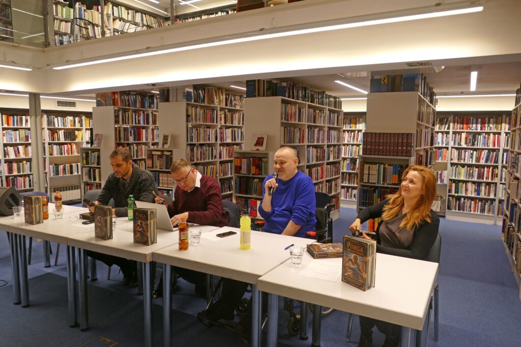 Panel-rasprava u knjižnici s policama punim knjiga, četiri sudionika sjede za stolom s mikrofonima i knjigama na stolu, jedan od njih govori u mikrofon.