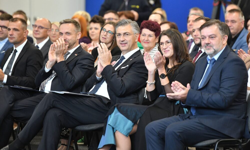 Na fotografiji je prikazana skupina ljudi koji sjede u redovima, očito prisustvujući formalnom događaju poput konferencije, prezentacije ili svečanosti. Većina sudionika odjevena je u poslovnu odjeću – odijela, haljine i elegantne kombinacije. Nekoliko osoba u prvom redu plješće, što sugerira da reagiraju na govor, izlaganje ili nastup. Pozadina je plava, a prostor djeluje profesionalno i organizirano.