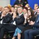 Na fotografiji je prikazana skupina ljudi koji sjede u redovima, očito prisustvujući formalnom događaju poput konferencije, prezentacije ili svečanosti. Većina sudionika odjevena je u poslovnu odjeću – odijela, haljine i elegantne kombinacije. Nekoliko osoba u prvom redu plješće, što sugerira da reagiraju na govor, izlaganje ili nastup. Pozadina je plava, a prostor djeluje profesionalno i organizirano.
