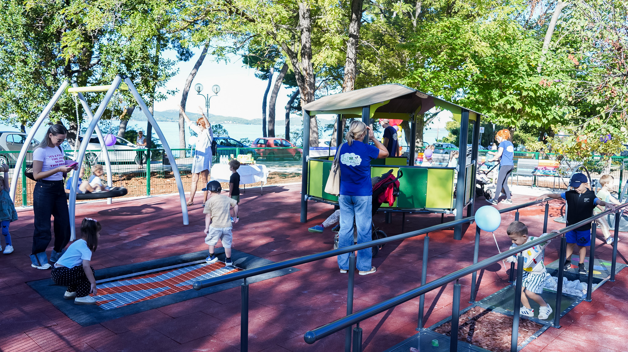 Na fotografiji se vidi novo senzorno dječje igralište uz more, okruženo zelenilom i drvećem. Djeca se igraju na različitim spravama – neka skaču po trampolinu, druga prolaze stazom od različitih materijala bosih nogu, dok se jedno dijete ljulja na velikoj ljuljački. U sredini igrališta nalazi se mala kućica s krovom, oko koje se također okupljaju mališani. Odrasli, među kojima su roditelji i odgojitelji, prate djecu, razgovaraju i fotografiraju. Atmosfera je vesela i opuštena, a u pozadini se vidi more i parkirani automobili uz šetnicu.