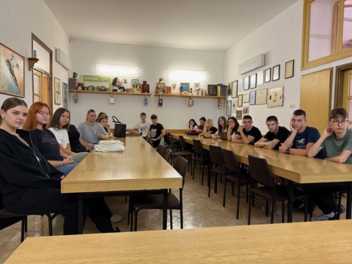 Na fotografiji se nalazi skupina mladih osoba koje sjede oko dva duga drvena stola u prostoriji koja izgleda kao učionica ili dvorana za sastanke. Zidovi su bijeli i ukrašeni raznim uokvirenim certifikatima, fotografijama i umjetničkim radovima. Na policama se nalaze knjige i dekorativni predmeti, a na kraju prostorije vidi se govornica. Osobe na slici djeluju pažljivo, kao da slušaju nekoga ili poziraju za zajedničku fotografiju. Cijela atmosfera sugerira obrazovno ili društveno okruženje, možda škola, kulturni centar ili mjesto za radionice i rasprave.