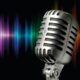 Na fotografiji se nalazi vintage mikrofon u prvom planu, s klasičnim metalnim dizajnom i rešetkastom glavom, postavljen na stalku. Iza mikrofona proteže se šareni uzorak zvučnog vala, koji prikazuje spektar boja — od ljubičaste i plave, preko zelene i žute, sve do crvene. Taj vizualni prikaz sugerira prisutnost zvuka, glazbe ili snimanja.