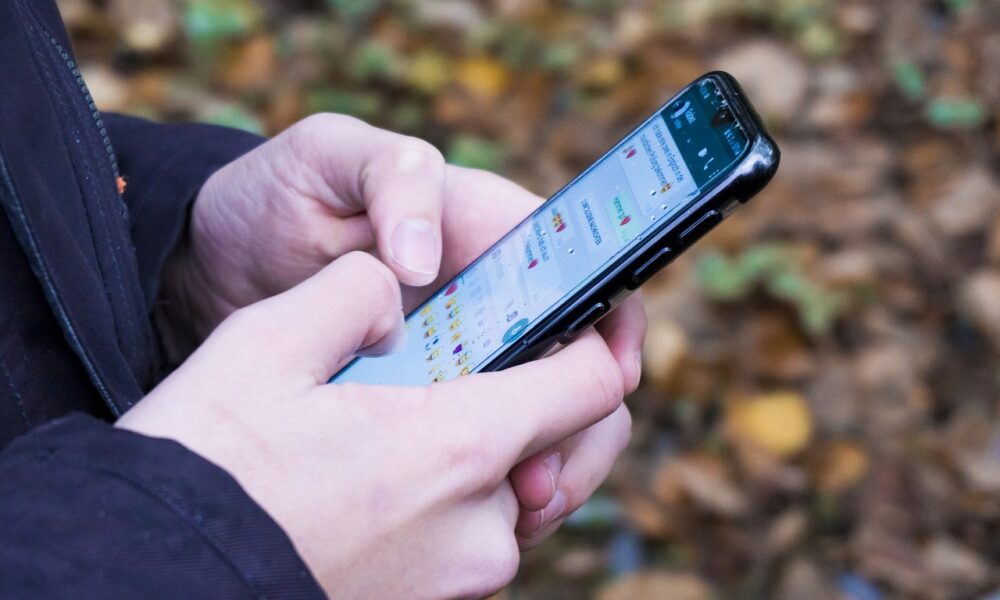 Na fotografiji se vidi osoba koja drži pametni telefon u rukama i tipka poruke. Na ekranu je otvorena aplikacija za dopisivanje, a razgovori sadrže puno emoji simbola. U pozadini se na tlu nalaze smeđi i žuti listovi, što sugerira da je fotografija snimljena u jesen ili u prirodi. Fokus je na rukama i mobitelu, dok je pozadina blago zamućena.