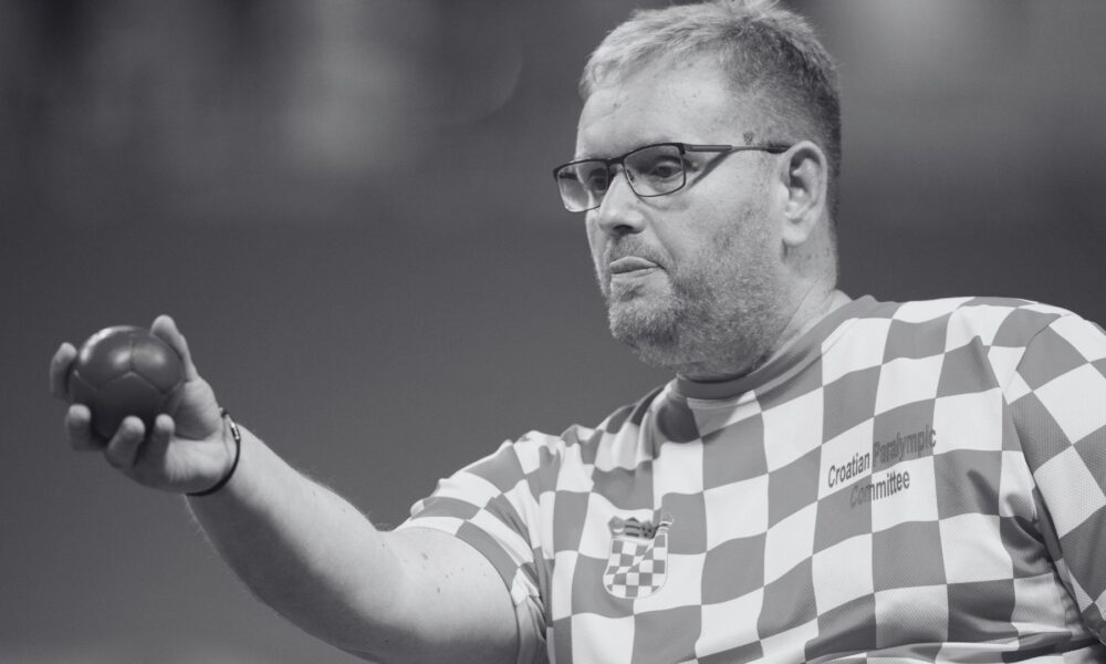 Crno-bijela fotografija hrvatskog boćara u dresu s kvadratićima, s natpisom „Croatian Paralympic Committee“. Sportaš nosi naočale i drži boću u ruci, koncentrirano gledajući prema naprijed.