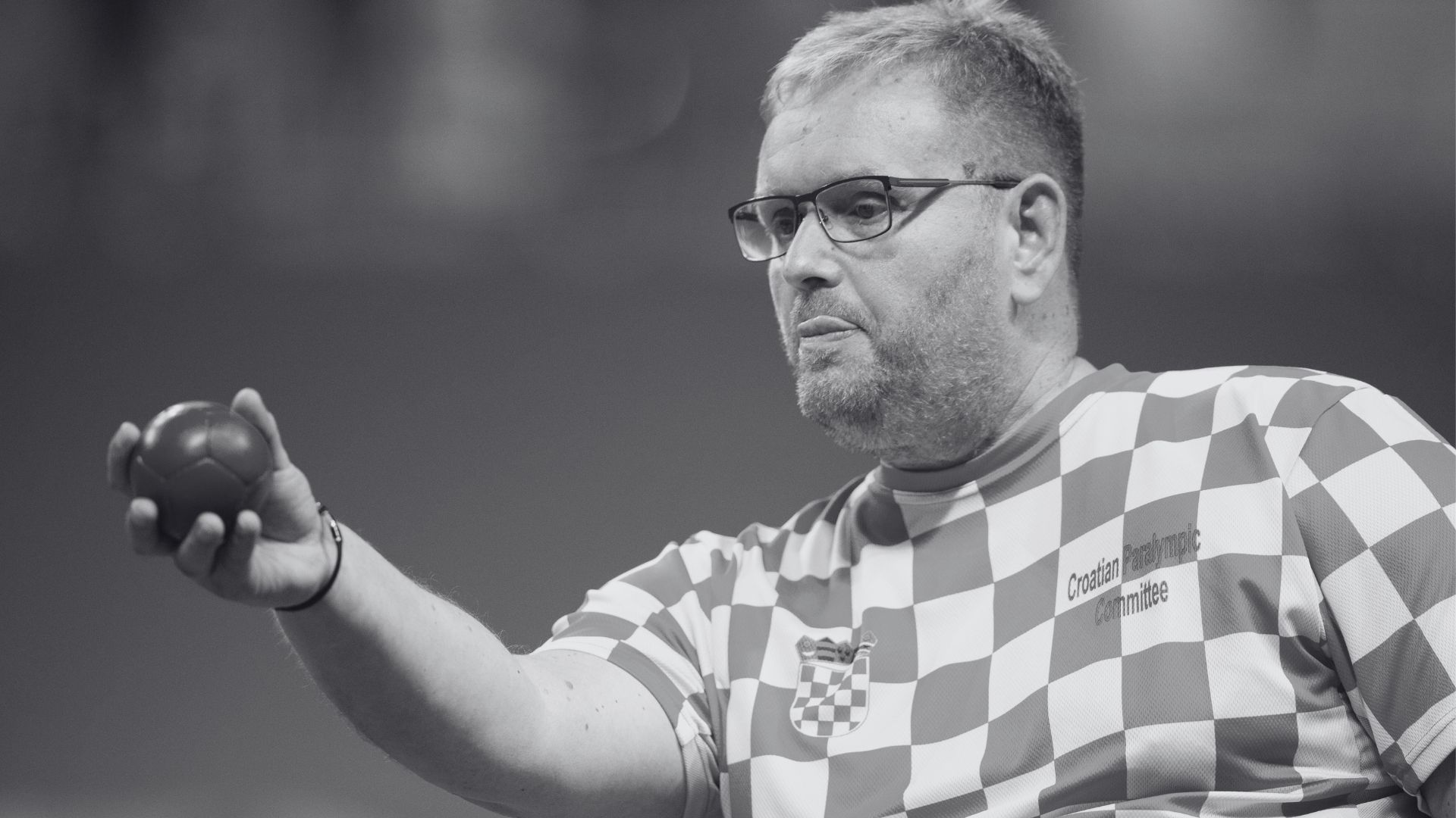 Crno-bijela fotografija hrvatskog boćara u dresu s kvadratićima, s natpisom „Croatian Paralympic Committee“. Sportaš nosi naočale i drži boću u ruci, koncentrirano gledajući prema naprijed.