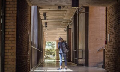 Na fotografiji se nalazi osoba koja stoji u hodniku zgrade, vjerojatno škole ili fakulteta. Hodnik ima zidove od crvene cigle s obje strane i pod prekriven pločicama. Strop je otvoren, s vidljivim gredama i rasvjetnim tijelima. Osoba je odjevena ležerno, nosi ruksak i gleda u mobitel ili neki uređaj u rukama. Sunčeva svjetlost dolazi kroz prozore s lijeve strane, stvarajući jake sjene i osvjetljavajući mjesto gdje osoba stoji. Na kraju hodnika vidi se otvoreniji prostor s drvećem, što sugerira da se izvan zgrade nalazi dvorište ili vrt.