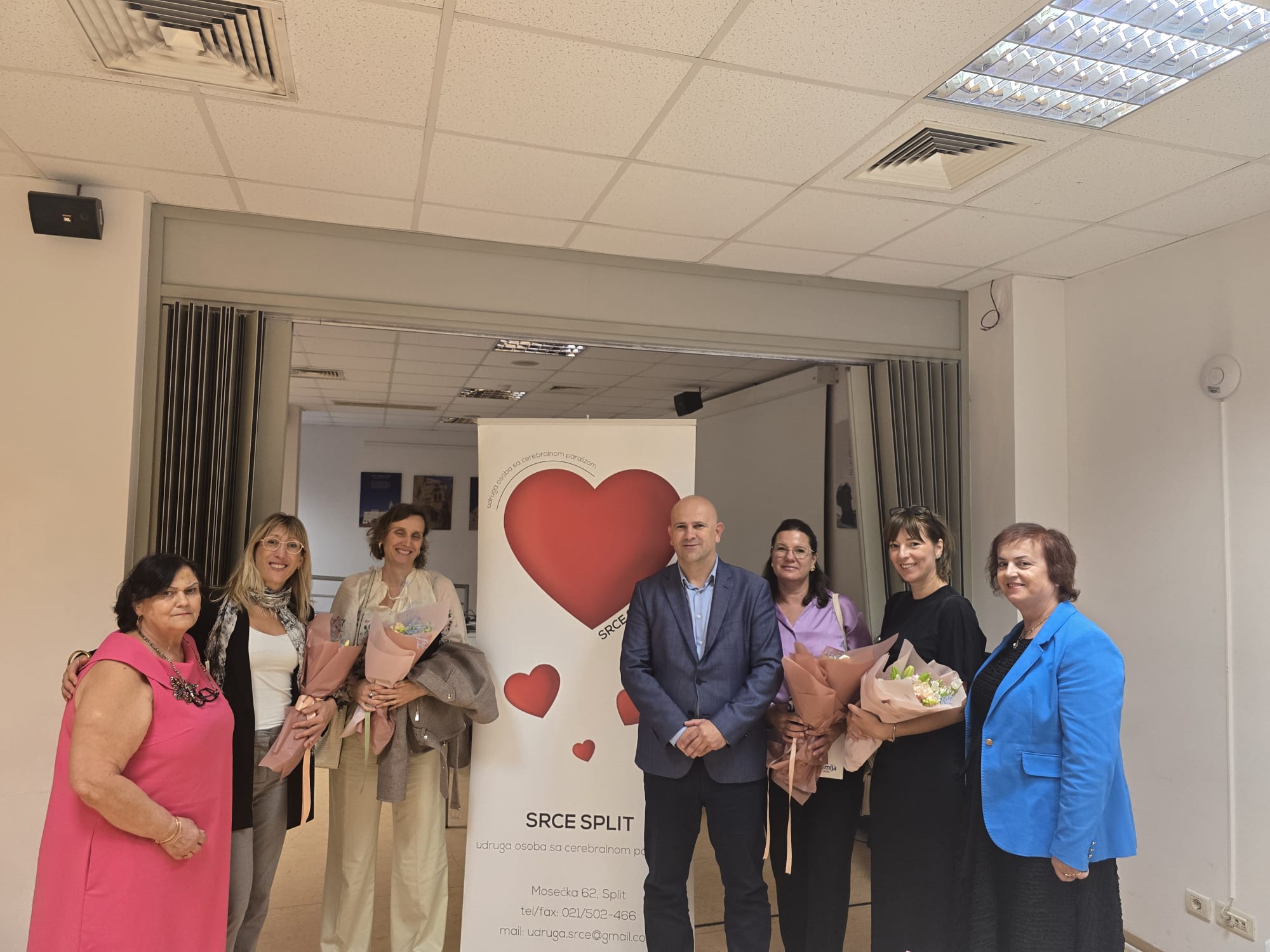 Na fotografiji je sedam odraslih osoba koje stoje u zatvorenom prostoru ispred bannera s velikim crvenim srcem i tekstom udruge "Srce Split – Udruga osoba sa cerebralnom paralizom". Osobe su odjevene svečano ili polusvečano, a tri od njih drže bukete cvijeća, što sugerira da su možda nagrađeni, zahvaljeni ili posebno istaknuti u sklopu događaja.