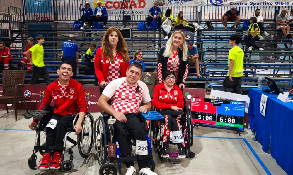 Slika prikazuje nekoliko ljudi na boccia terenu od kojih je troje u kolicima. Svi su obučeni u crvena hrvatska obilježja.