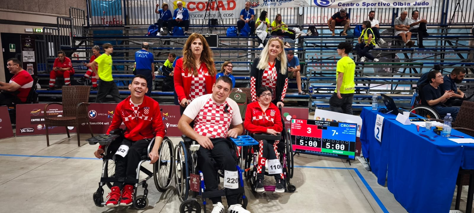 Slika prikazuje nekoliko ljudi na boccia terenu od kojih je troje u kolicima. Svi su obučeni u crvena hrvatska obilježja.