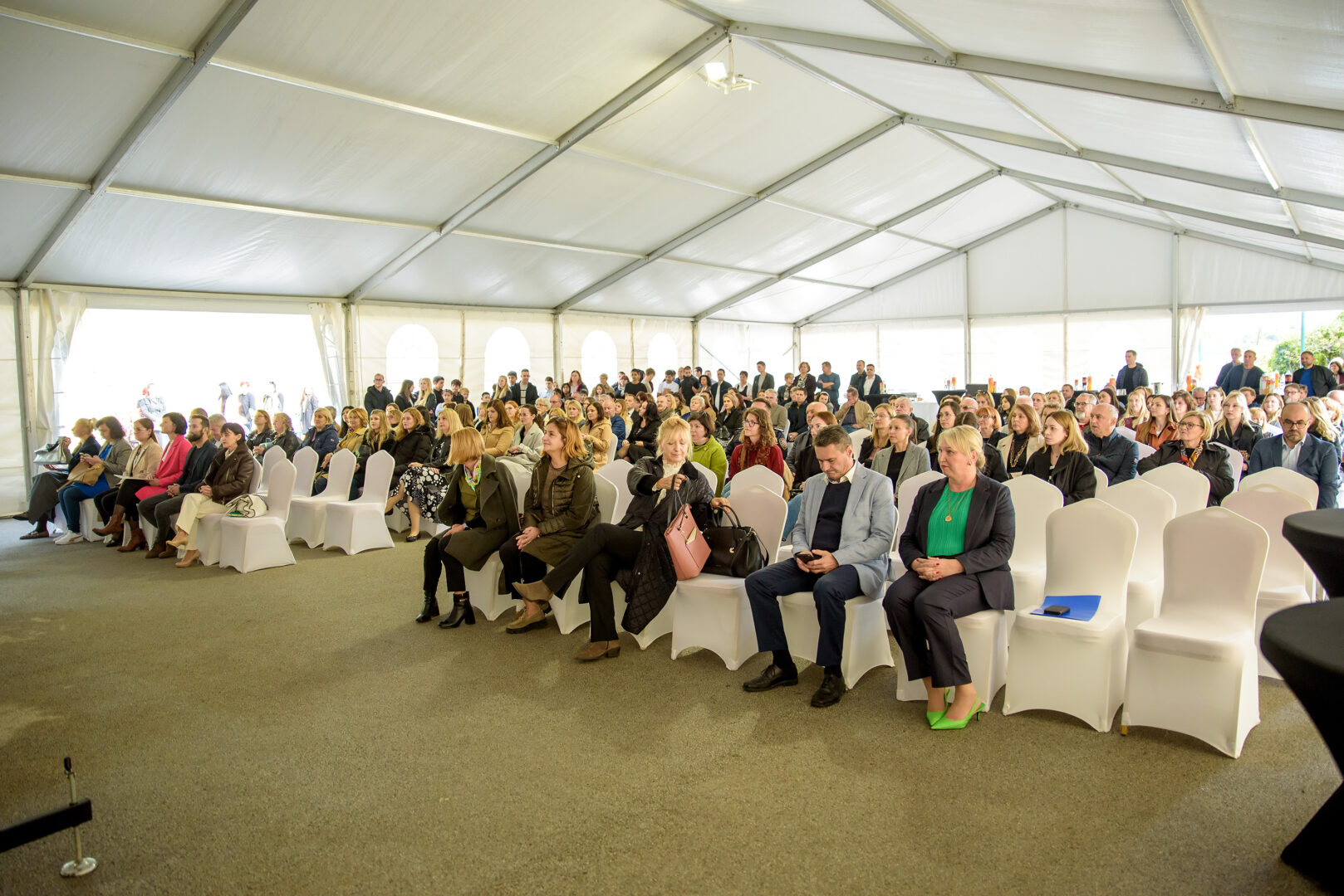 Na fotografiji je prikazan veliki broj ljudi okupljenih unutar prostranog bijelog šatora, koji djeluje kao mjesto za svečani ili zajednički događaj. Stolice su uredno raspoređene u redove i prekrivene bijelim navlakama, što prostoru daje svečan i organiziran izgled. Prirodna svjetlost prodire kroz prozirne stranice šatora, stvarajući ugodnu i otvorenu atmosferu. Publika je raznolika — neki su odjeveni formalno, drugi ležernije, a u prednjem redu ima nekoliko praznih mjesta. Dio prisutnih razgovara, dok drugi gledaju u svoje telefone, što sugerira da se još čeka početak programa. Iako nije vidljiva pozornica ni govornik, raspored i ambijent upućuju na događaj poput konferencije, seminara ili svečanosti u zajednici.