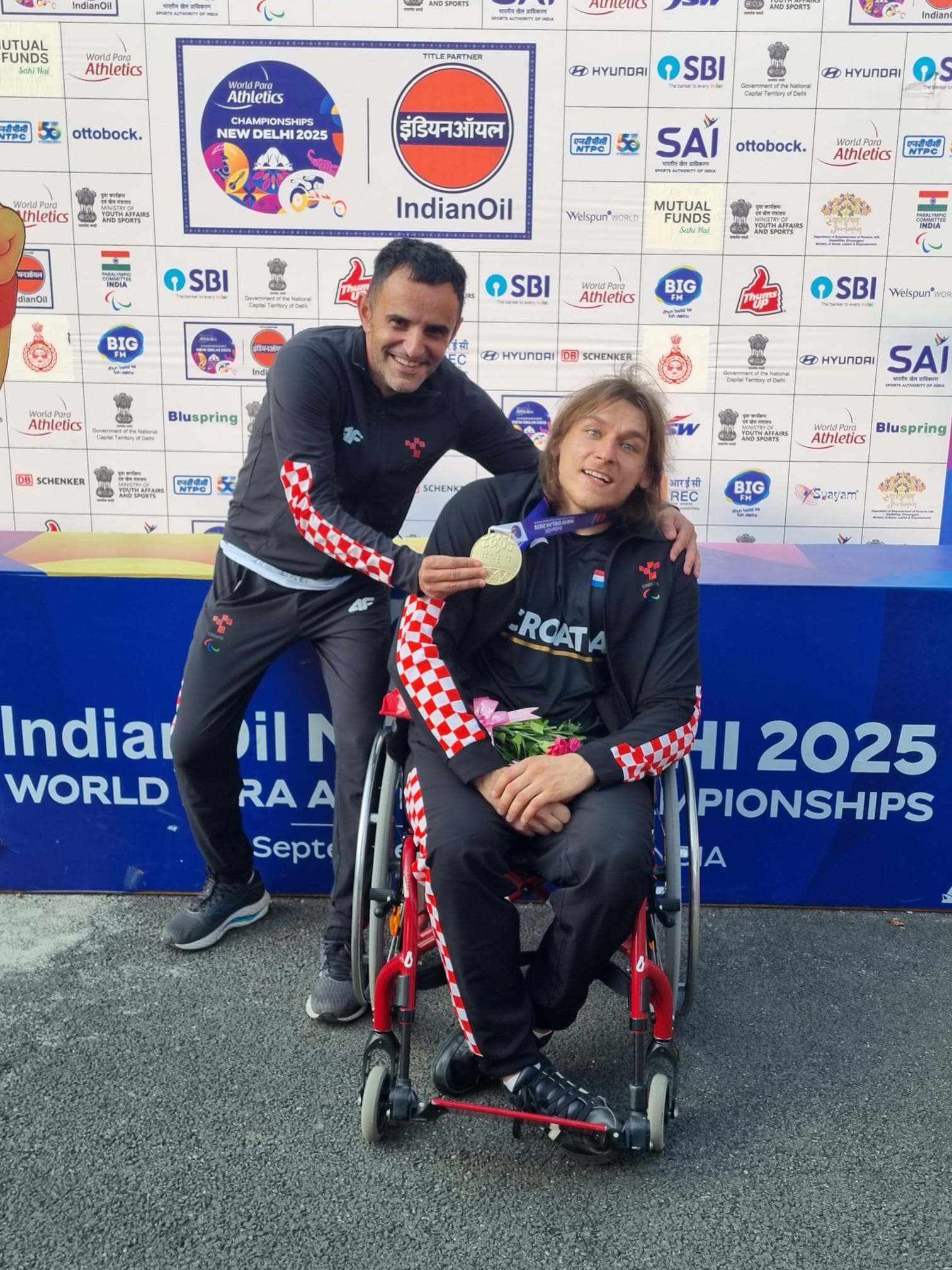 Fotografija prikazuje dva čovjeka. Jedan je u kolicima s medaljom oko vrata, a drugi ga drži za medalju i pozira s njim.