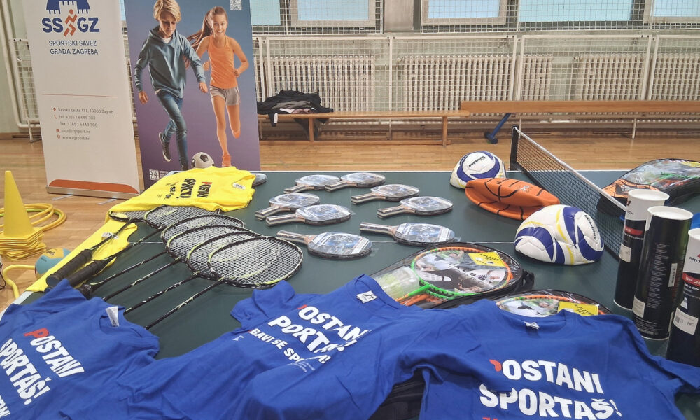 Na stolu u sportskoj dvorani izložena je sportska oprema – reketi, loptice, lopte i majice s natpisom „Postani sportaš“.