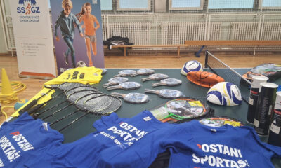 Na stolu u sportskoj dvorani izložena je sportska oprema – reketi, loptice, lopte i majice s natpisom „Postani sportaš“.
