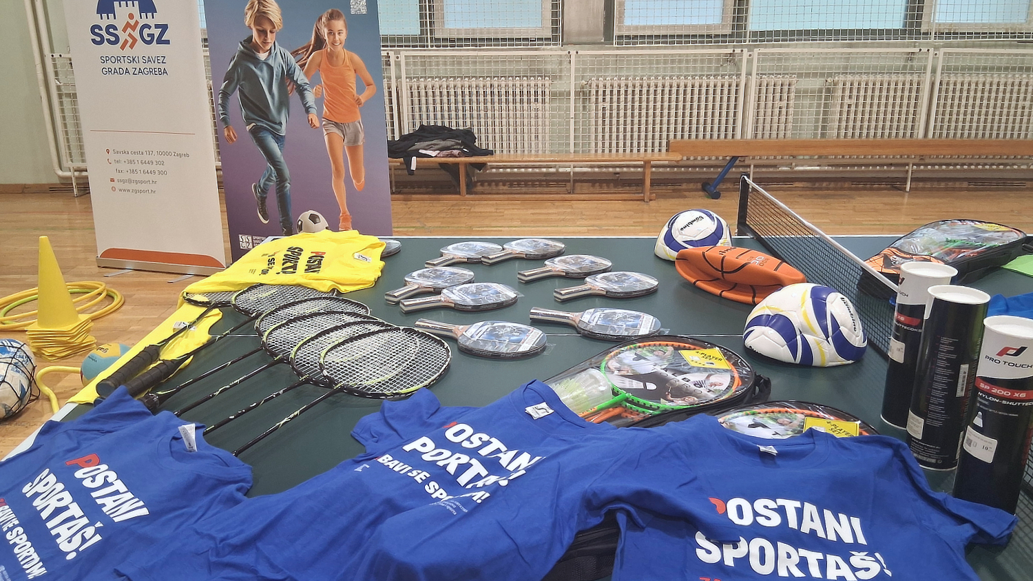 Na stolu u sportskoj dvorani izložena je sportska oprema – reketi, loptice, lopte i majice s natpisom „Postani sportaš“.