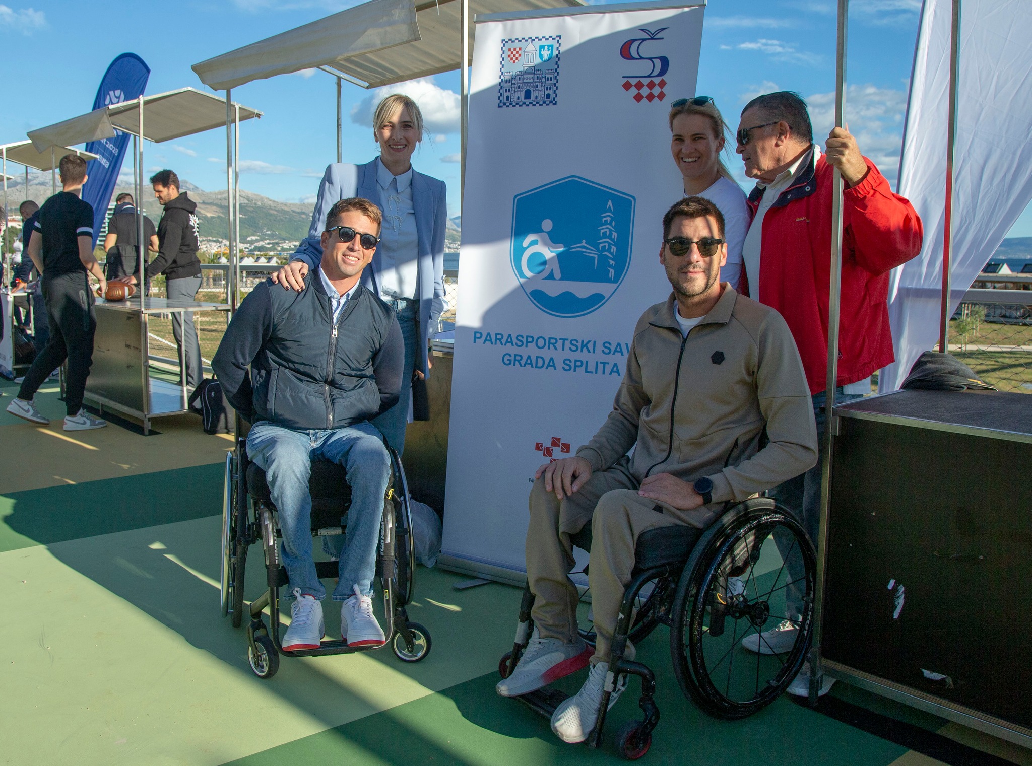 Fotografija prikazuje dva čovjeka u invalidskim kolicima, dok iza njih stoji troje ljudi te se nsalazi baner na kojem piše 'Parasportski savez grada Splita'.