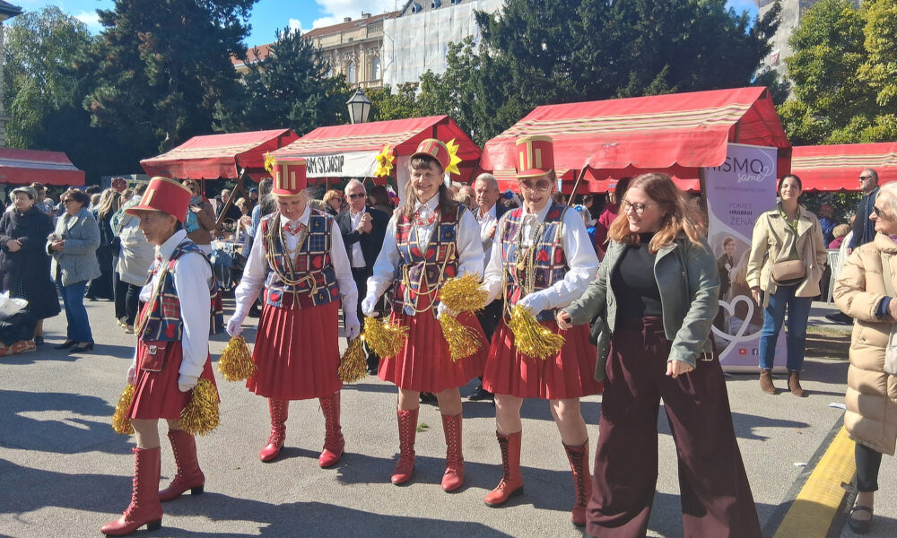 Skupina žena u šarenim kostimima i crvenim čizmama pleše na otvorenom tijekom manifestacije, okružena publikom.