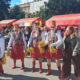Skupina žena u šarenim kostimima i crvenim čizmama pleše na otvorenom tijekom manifestacije, okružena publikom.
