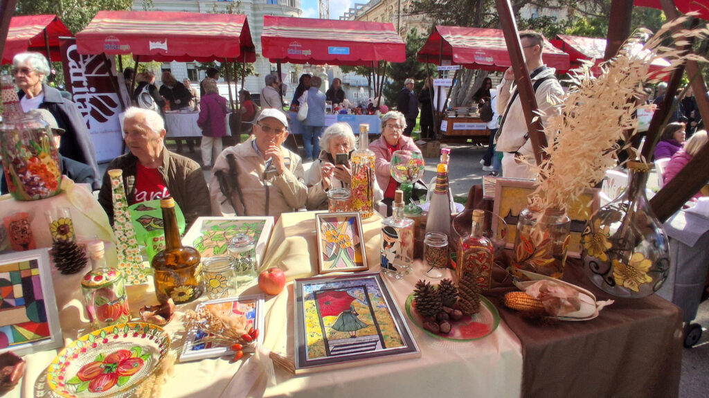Štand na manifestaciji s izloženim rukotvorinama i ukrasnim predmetima, dok nekoliko starijih osoba sjedi iza stola.