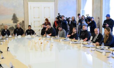 Fotografija prikazuje više ljudi koji sjede za stolom, a iza njih stoje kamermani i snimaju ih.
