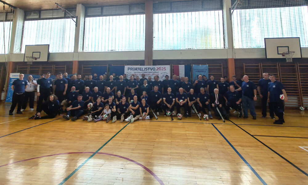 Velika skupina sudionika u sportskim majicama pozira u dvorani s košarkaškim koševima ispod transparenta „Prijateljstvo 2025“, s loptama za odbojku ispred sebe.