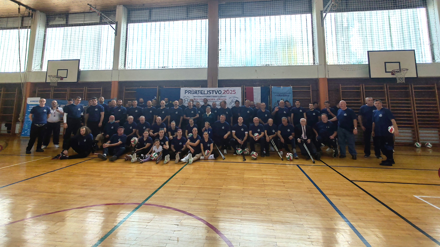 Velika skupina sudionika u sportskim majicama pozira u dvorani s košarkaškim koševima ispod transparenta „Prijateljstvo 2025“, s loptama za odbojku ispred sebe.