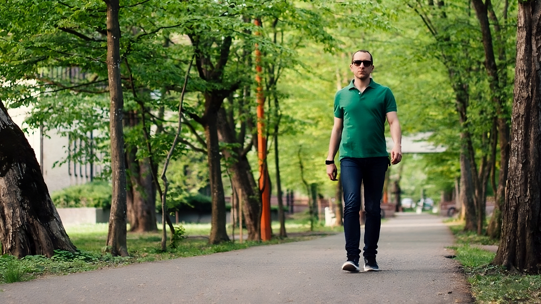 Muškarac u zelenoj majici i trapericama hoda parkom po stazi okruženoj drvećem, noseći sunčane naočale i pametni sat.