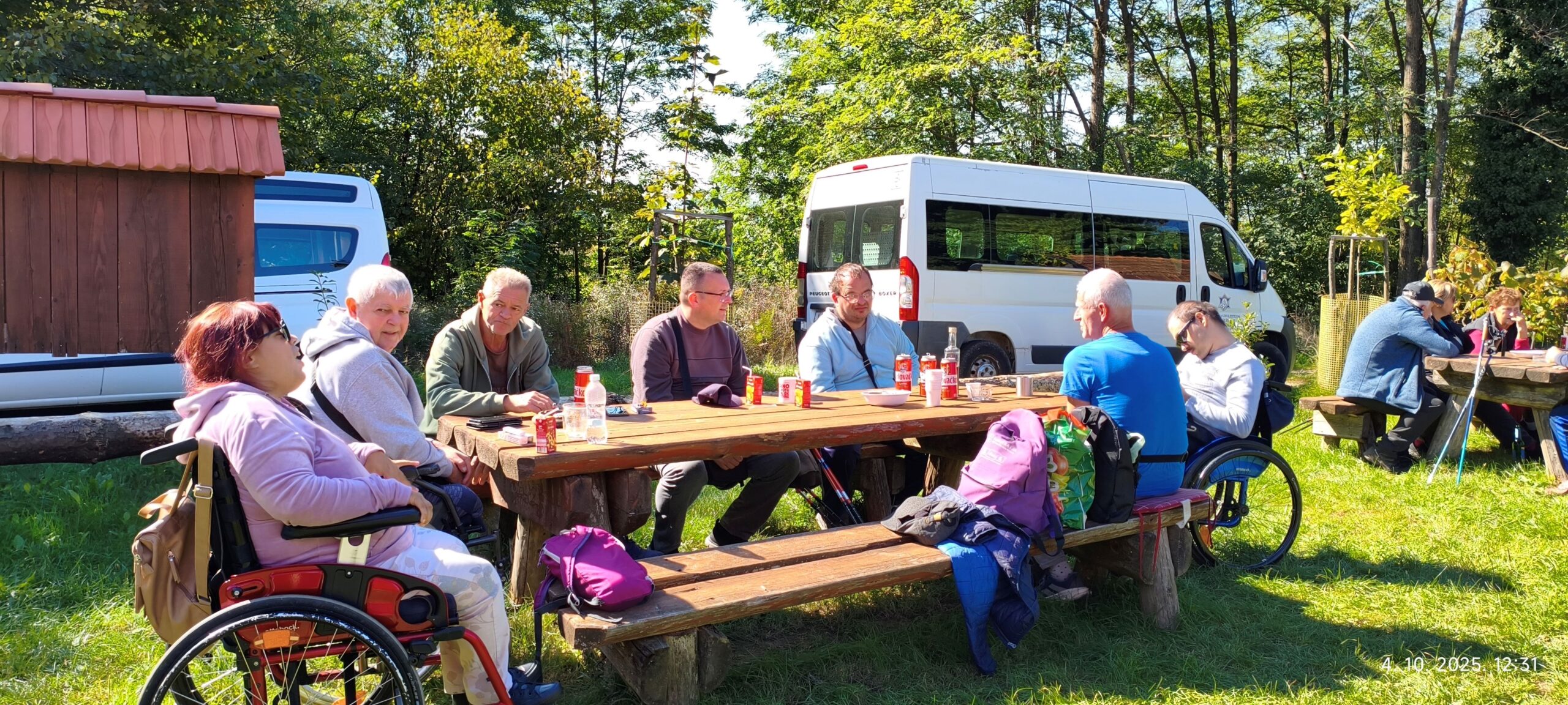 Na fotografiji je prikazana skupina ljudi okupljena na otvorenom prostoru, oko drvenih stolova za piknik, smještenih na travnatoj površini okruženoj drvećem. Nekoliko osoba sjedi, a neke su u invalidskim kolicima, što ukazuje na inkluzivno i pristupačno druženje. Ljudi razgovaraju i uživaju u obroku, a na stolovima se nalaze boce, šalice i torbe, što dodatno naglašava opuštenu i prijateljsku atmosferu. U pozadini se vide dva bijela kombija i drvena građevina, što sugerira organiziranu logistiku i moguće sadržaje za sudionike. Vrijeme je sunčano i ugodno, što doprinosi vedrom ozračju zajedništva.