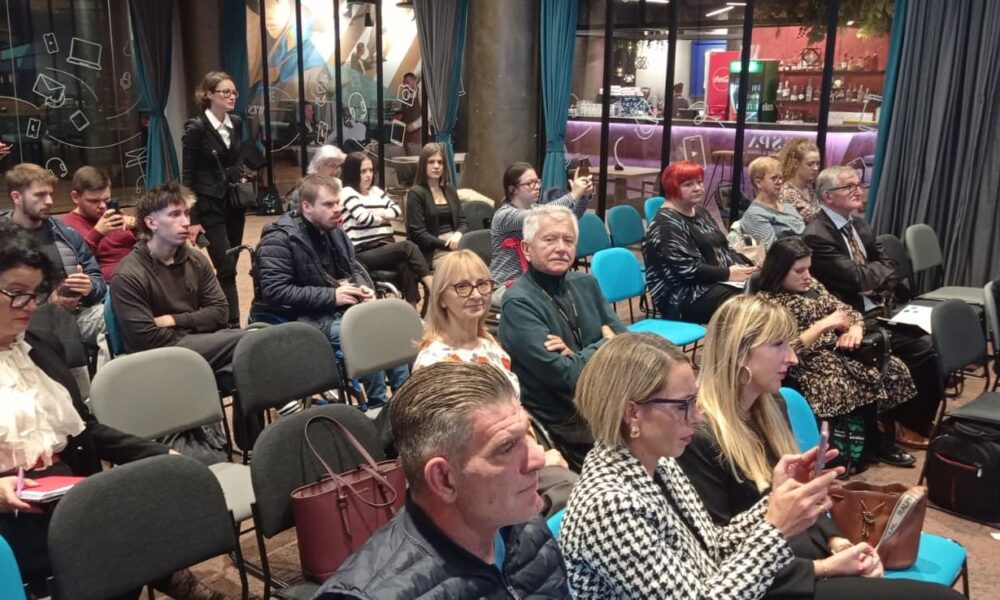 Fotografija prikazuje skupinu ljudi okupljenih u suvremeno uređenoj prostoriji, najvjerojatnije na predavanju, prezentaciji ili zajedničkom sastanku. Većina sudionika sjedi na šarenim stolicama raspoređenima u redove, dok nekoliko osoba stoji ili koristi mobilne telefone. U pozadini se vidi staklena pregrada koja otkriva svijetli prostor s stolovima i stolicama — moguće kafić, lounge ili zajednički prostor za odmor. Sudionici su različitih dobnih skupina, neki drže bilježnice ili torbe, što dodatno naglašava edukativni ili informativni karakter događanja. Atmosfera je mirna i koncentrirana, a prostor odiše otvorenošću i funkcionalnošću, pogodnom za razmjenu znanja i ideja.