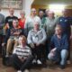 Fotografija prikazuje nekoliko ljudi kako stoje u zatvorenom prostoru. Neki od njih imaju medalje oko vrata, a jedna osoba je u invalidskim kolicima.