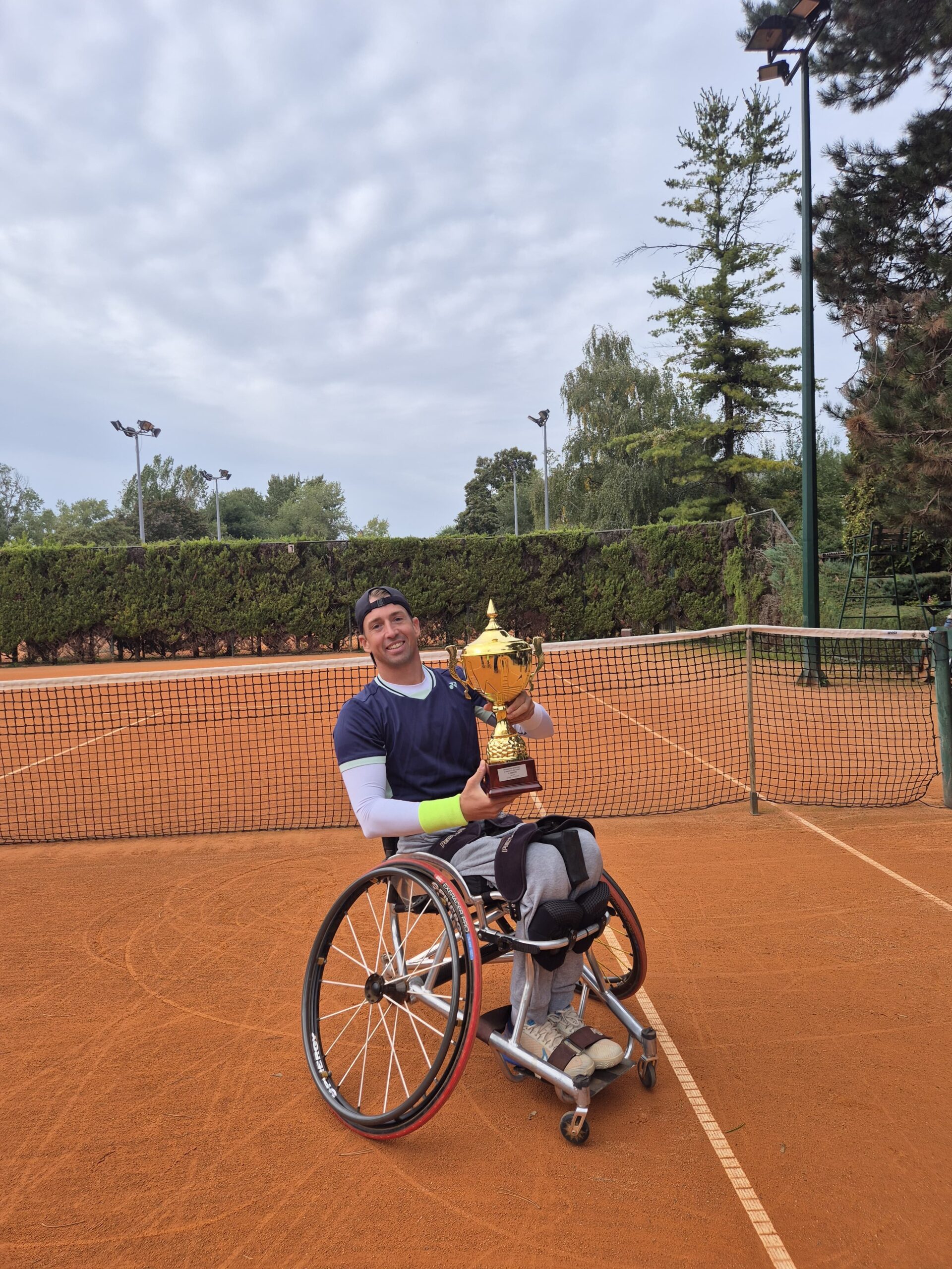 Na fotografiji je tenisač u kolicima koji drži pehar, a nalazi se na teniskom terenu.