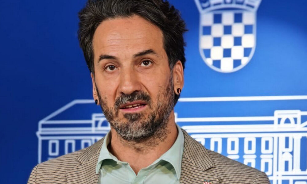 Muškarac govori ispred plavog zida s hrvatskim grbom, odjeven u sako i svijetlu košulju, tijekom službenog obraćanja medijima.