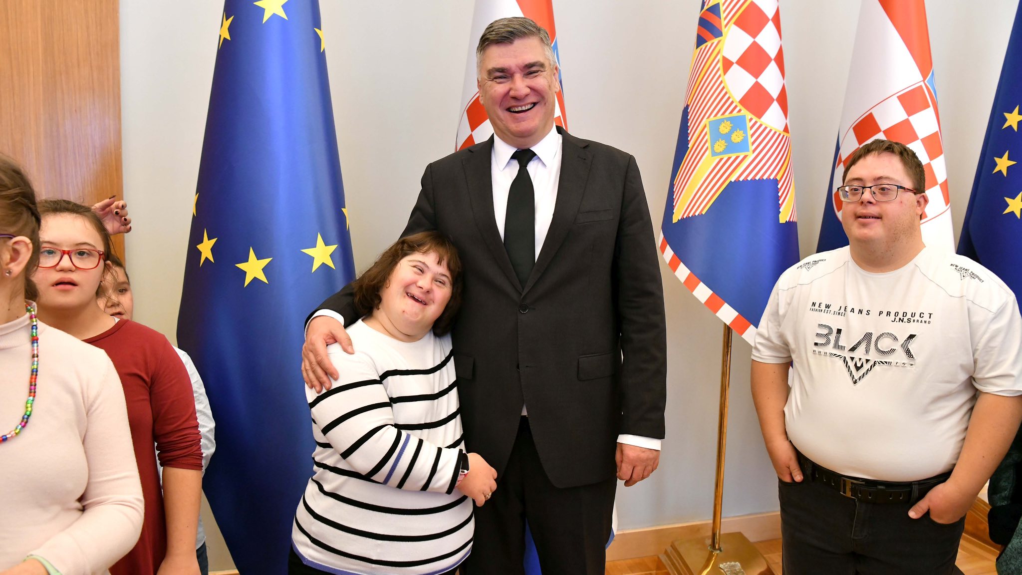 Na fotografiji je čovjek u odjelu koji krli djevojčicu sa Down Sindromom, a oko njega su i druga djeca s Down sindromom. Iza njih je nekoliko zastava, a između ostalog hrvatska zastava i zastava Europske Unije.