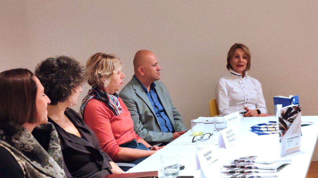 Panel-rasprava u galerijskom prostoru – više osoba sjedi za stolom s knjigama i oznakama s imenima, dok jedna sudionica govori i gestikulira tijekom predstavljanja publikacije.