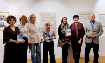 Sudionici svečanosti stoje u nizu u galerijskom prostoru ispred izložbenih panoa; nekoliko osoba drži plakete i nagrade u rukama, a svi se smiješe prema kameri.