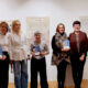 Sudionici svečanosti stoje u nizu u galerijskom prostoru ispred izložbenih panoa; nekoliko osoba drži plakete i nagrade u rukama, a svi se smiješe prema kameri.
