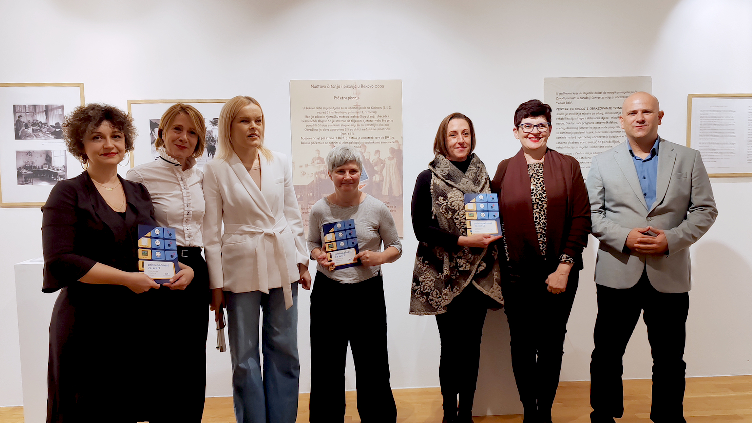 Sudionici svečanosti stoje u nizu u galerijskom prostoru ispred izložbenih panoa; nekoliko osoba drži plakete i nagrade u rukama, a svi se smiješe prema kameri.