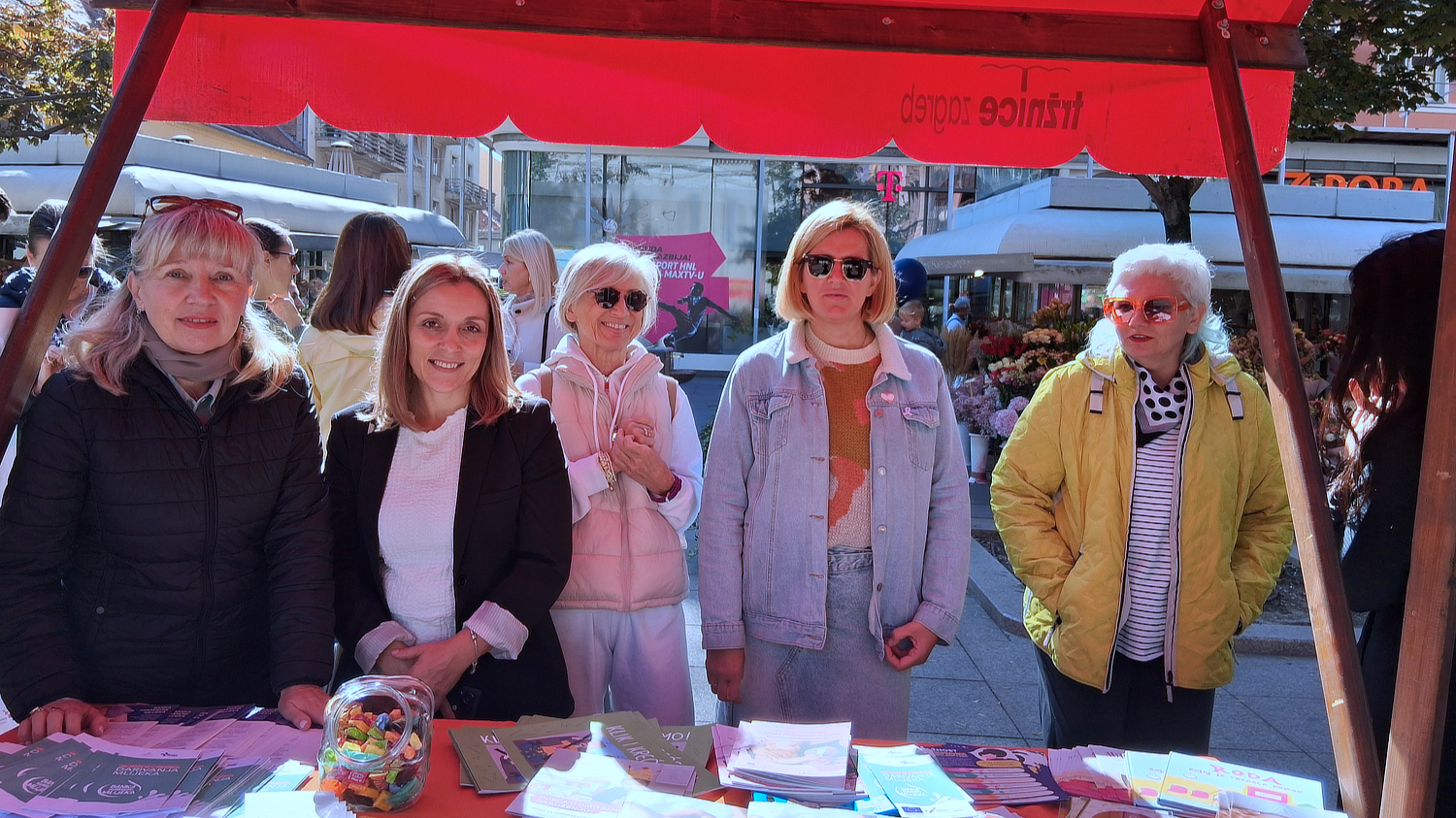 Skupina žena stoji iza štanda prekrivenog informativnim materijalima i promotivnim letcima tijekom obilježavanja Nacionalnog tjedna dojenja.