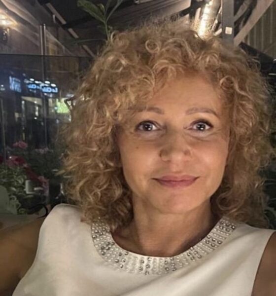 Na fotografiji je osoba svijetle puti s kovrčavom plavom kosom, odjevena u bijeli gornji dio ukrašen dekorativnim elementima oko izreza. Pozadina je slabo osvijetljen prostor, moguće interijer ili eksterijer s reflektirajućim površinama i vidljivim biljkama. Atmosfera djeluje intimno i pomalo svečano, s blagim kontrastom između osobe u prvom planu i zamagljenog ambijenta iza nje.