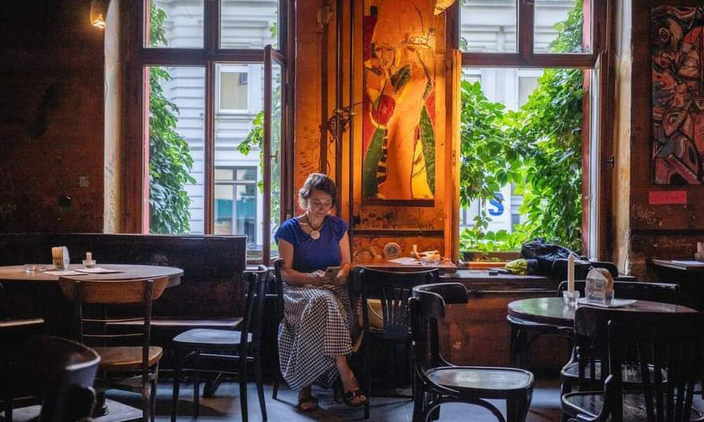 Žena sjedi sama u umjetnički uređenom kafiću uz veliko prozorsko okno obraslo zelenilom, dok svjetlost izvana obasjava prostor i stvara mirnu, intimnu atmosferu.