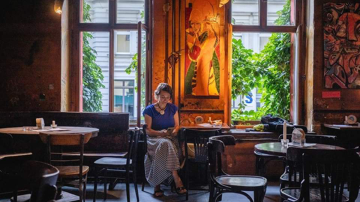 Žena sjedi sama u umjetnički uređenom kafiću uz veliko prozorsko okno obraslo zelenilom, dok svjetlost izvana obasjava prostor i stvara mirnu, intimnu atmosferu.