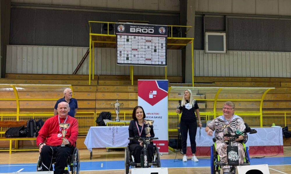 Fotografija prikazuje troje ljudi u invalidskim kolicima kako stoje iza broja mjesta kojeg su osvojili na sportskom natjecanju. Osoba u sredini drži pehar i nosi zlatnu medalju oko vrata, dok druga dvojica s njezine ljeve i desne strane oko vrata nose samo medalje, jedan srebrnu, drugi brončanu.