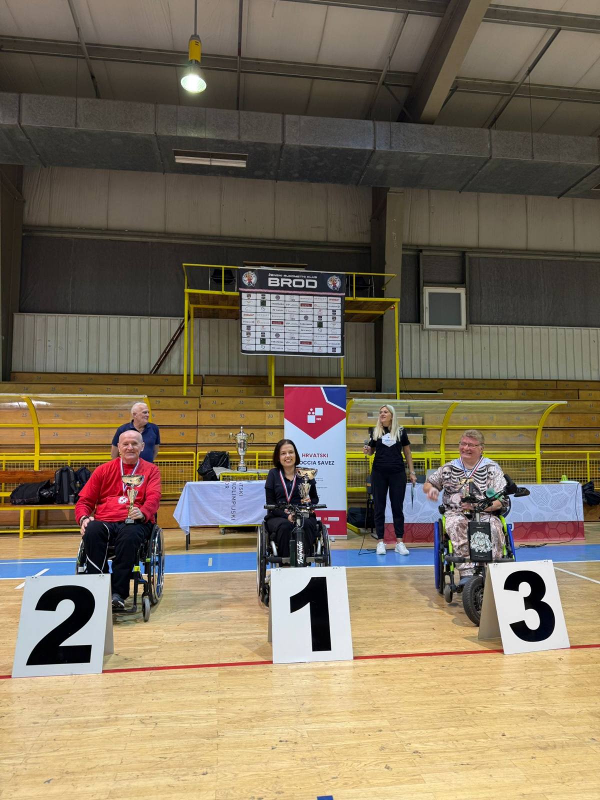 Fotografija prikazuje troje ljudi u invalidskim kolicima kako stoje iza broja mjesta kojeg su osvojili na sportskom natjecanju. Osoba u sredini drži pehar i nosi zlatnu medalju oko vrata, dok druga dvojica s njezine ljeve i desne strane oko vrata nose samo medalje, jedan srebrnu, drugi brončanu.