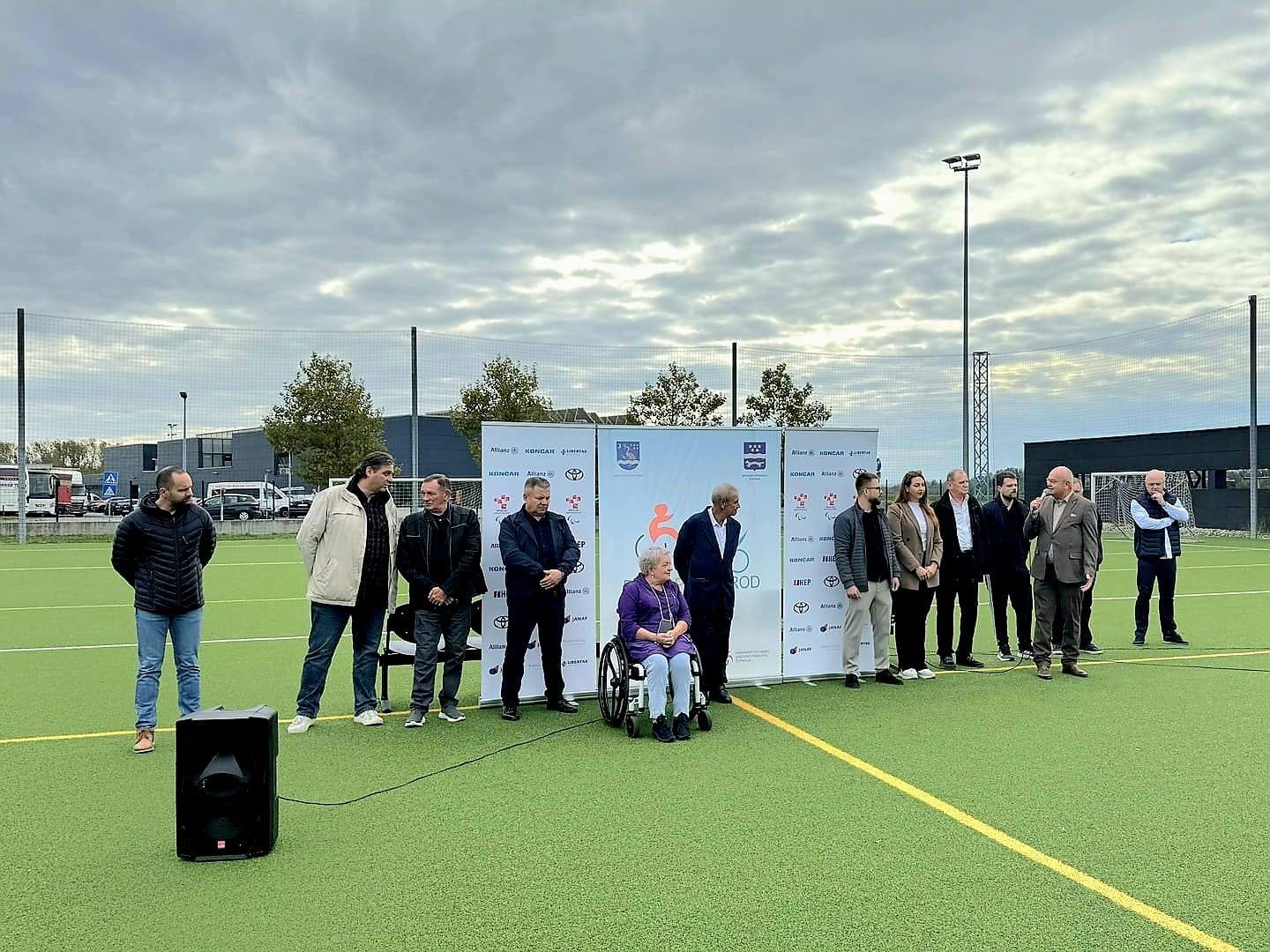 Fotografija prikazuje nekoliko ljudi lijepoi obučenih na sportskom igralištu. Jedna osoba je u invalidskim kolicima, dok drugi stoje.