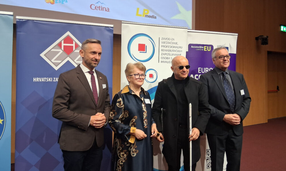 Četvero sudionika u formalnoj odjeći stoji ispred promotivnih panoa Hrvatskog zavoda za zapošljavanje, Zavoda za vještačenje i EU Accessible Centra, tijekom simpozija osoba s invaliditetom.