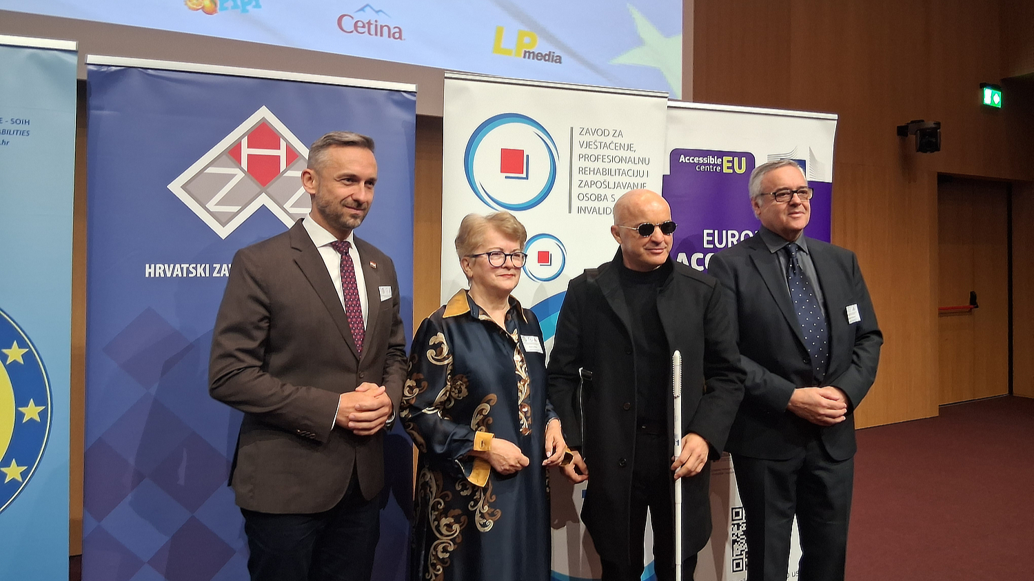 Četvero sudionika u formalnoj odjeći stoji ispred promotivnih panoa Hrvatskog zavoda za zapošljavanje, Zavoda za vještačenje i EU Accessible Centra, tijekom simpozija osoba s invaliditetom.