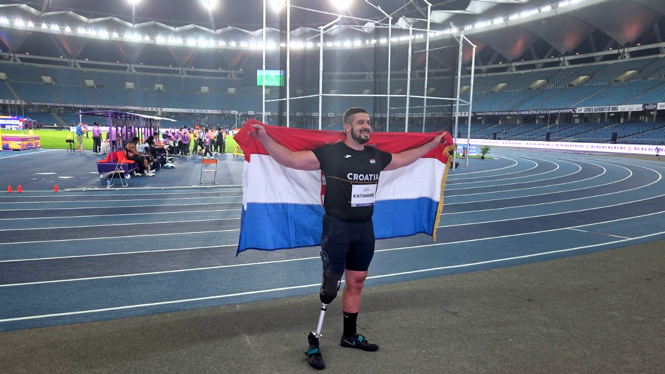 Atletičar s protezom na nozi stoji na atletskoj stazi i s osmijehom drži hrvatsku zastavu raširenu iza leđa, nakon osvajanja medalje na natjecanju.